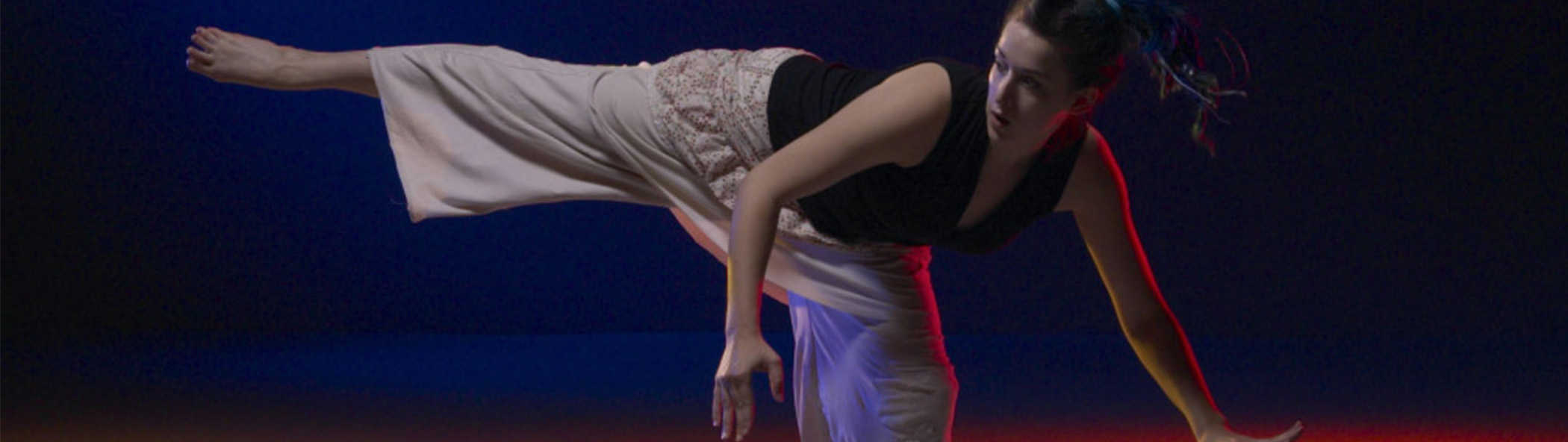 A dancer, Julia, in cream colored pants and black shirt. Extends her right leg out laterally and counter balances by leaning her upper body over to the other side. She’s lit by red and white lighting and a dark blue backdrop lies in the background.