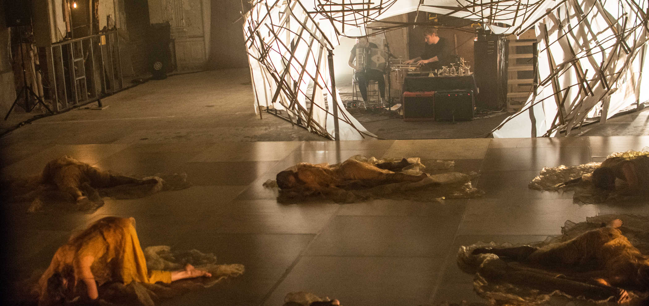 Bodies riddled across a Marley stage, all dressed in beige and earth tone fabrics. Many are lying face down on the floor, others in fetal position or child’s pose. In the background is a white tented structure with stick-like beams. Underneath the structure sits two musicians playing drums and a piano accordion.