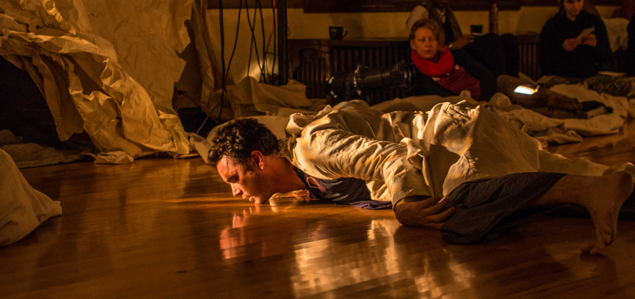 Performer Jonathan Meyer in a white coat, lies chin to the floor, face down. His knees are bent and his toes are tucked, as if he could propel himself forward by his toes at any time. Beyond Jonathan sits a couple of audience members, and more white fabric or tarp.