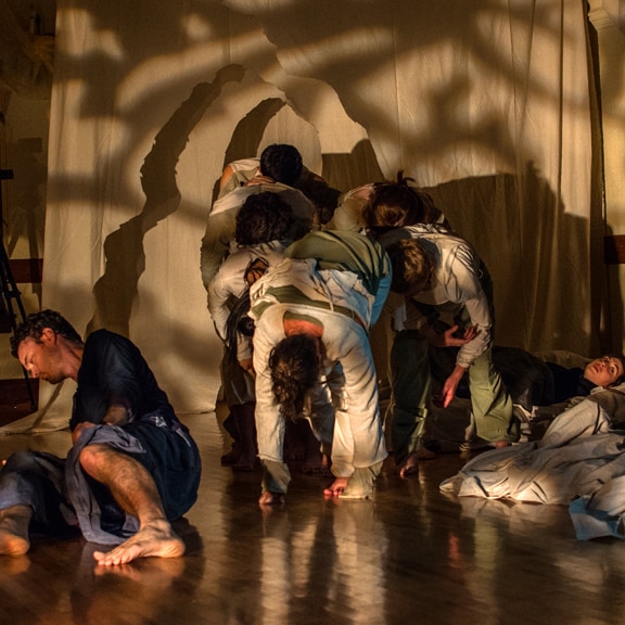 A group of dancers, mainly in green and white long sleeved shirts and pants, make a standing pile, heads stacked on each others mid to low backs. One other dancer, Jonathan Meyer, dressed in blue, lays just in front of the standing dancers, while Julia Antonick, another dancer, lays just behind the group of standing dancers. In the background is a beige backdrop full of shadows.