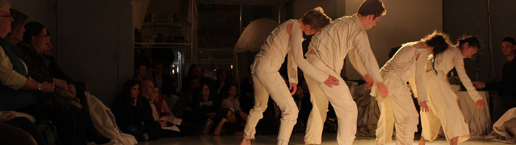Four dancers, dressed in white long sleeve shirts and long pants, walk in a line, each of them partnered up with the partner in the back resting their forehead on their partner’s back. The dancers perform on a white Marley stage surround by seated audience members on all sides.