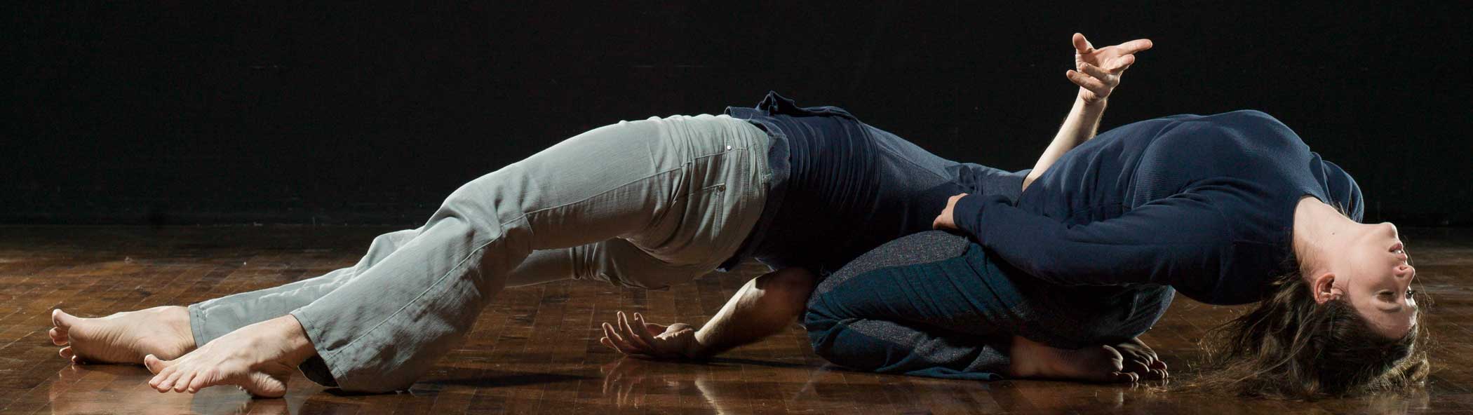 Dancers, Julia Antonick and Jonathan Meyer, perform on a floor both in arched back positions. Julia sits on her knees with her head and torso tossed back to the floor, while Jonathan, suspends part of his weight across Julia’s lap and arches the rest of his body off the ground, resting the rest of his weight only on his head and his heels. Both wear navy blue shirts and gray-toned pants.