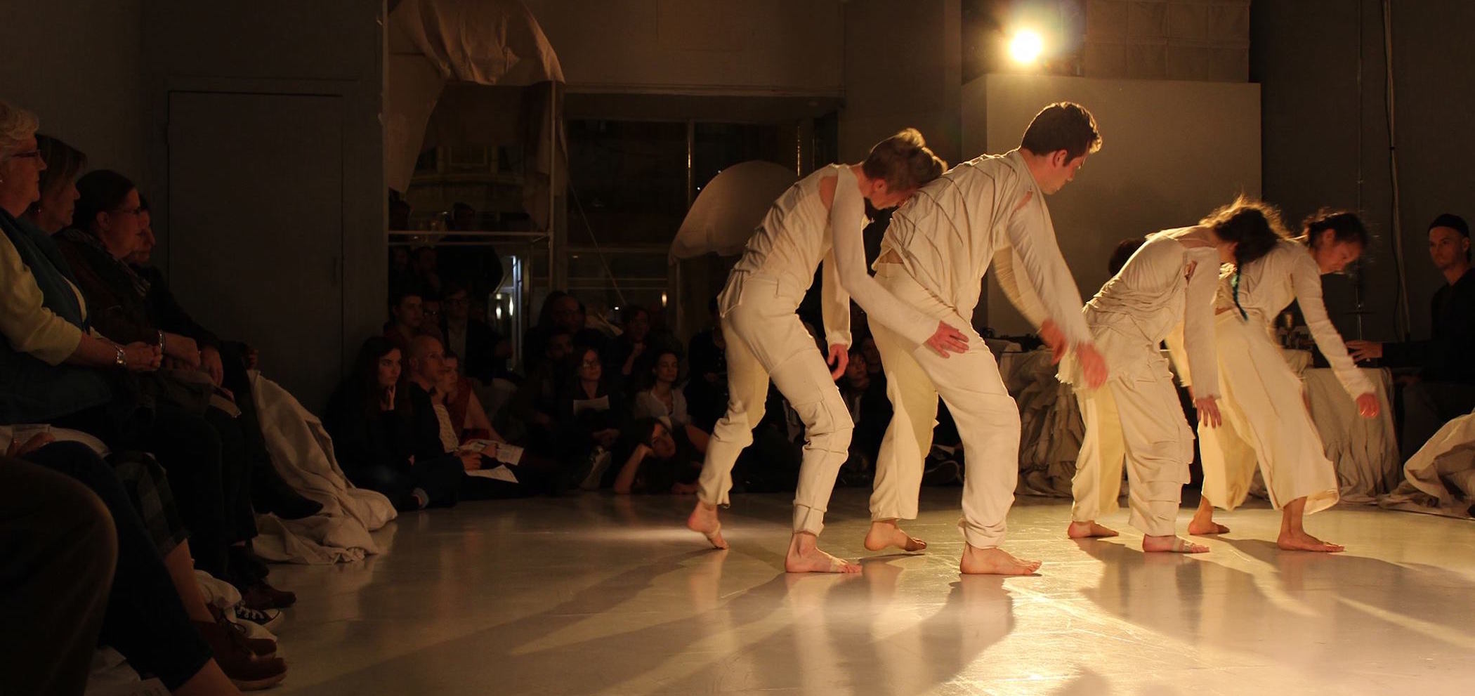 Four dancers, dressed in white long sleeve shirts and long pants, walk in a line, each of them partnered up with the partner in the back resting their forehead on their partner’s back. The dancers perform on a white Marley stage surround by seated audience members on all sides.