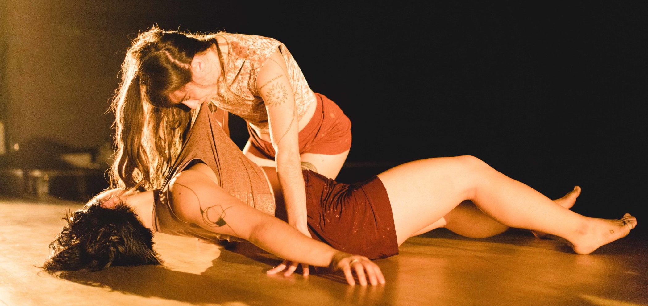 Two female dancers, both in shorts and sleeveless cream colored crop tops, perform on a yellow tinted lit stage. One dancer’s upper back is suspended just off the ground while the other dancer hovers over her with her dance partner’s shirt in her mouth, as if she’s holding her up by her teeth.