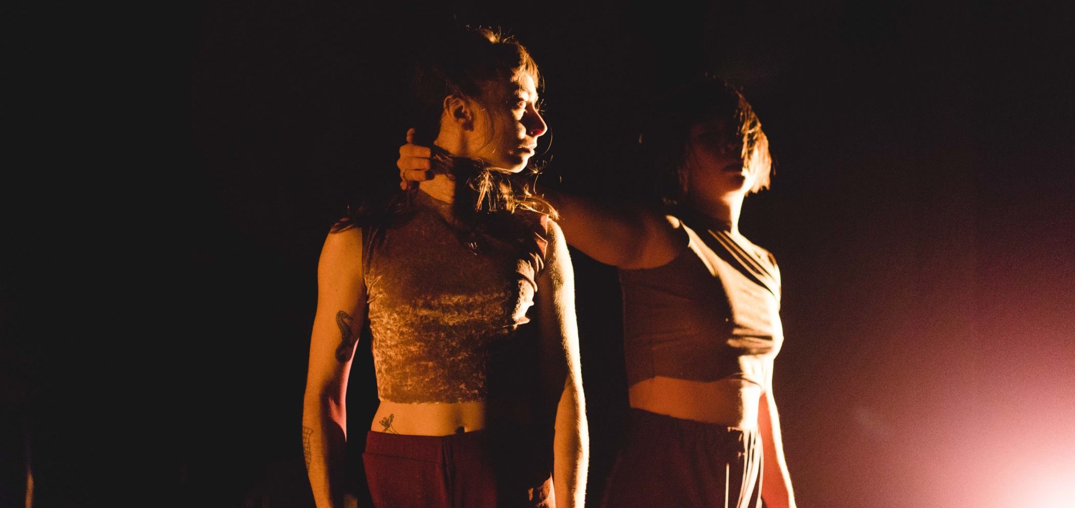 Two female dancers, both in shorts and sleeveless cream colored crop tops stand on a stage, illuminated only by a pink light on the bottom right corner. One dancer has her hand wrapped around the back of the neck of the other. Both stand tall, looking just past one another.