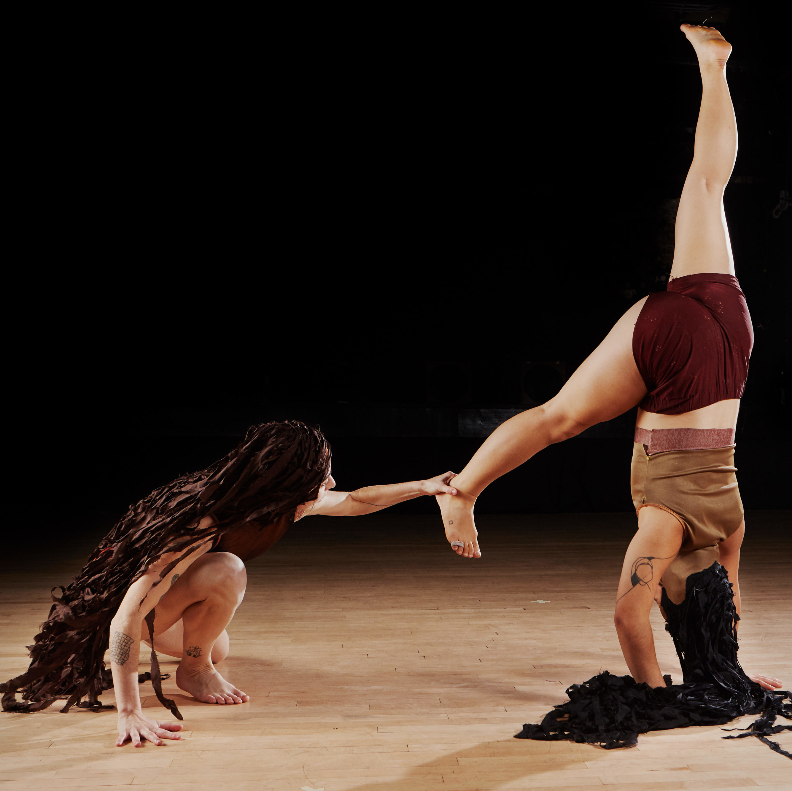 One female bodied dancer does a handstand while another female dancer pulls down her heel beneath her