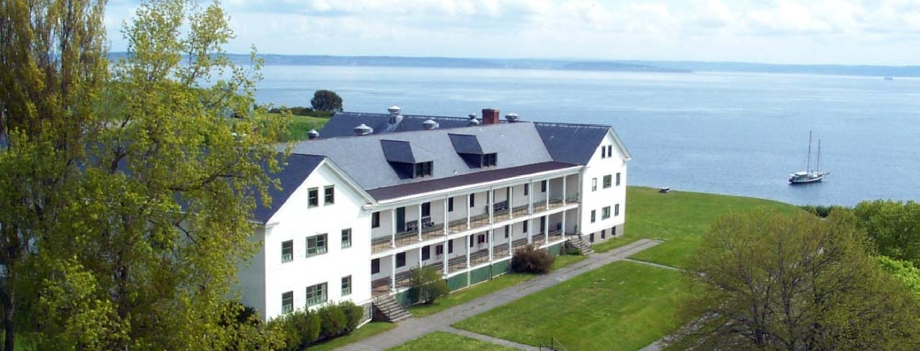 A big, white building laden with balconies overlooks a large body of water in Fort Worden, Washington. The foliage surrounding the building is green and lush, day is clear and bright. A sailboat sits just off shore.