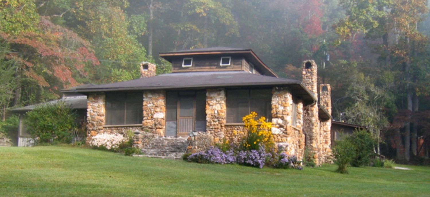 A stone house sits a top a grassy hill. A forest, just beginning to exhibit signs of autumn foliage, sits behind the house. Yellow and purple wild flowers line the outside of the stone house. The house is the Hambridge center for creative arts in Rabun County, Georgia.