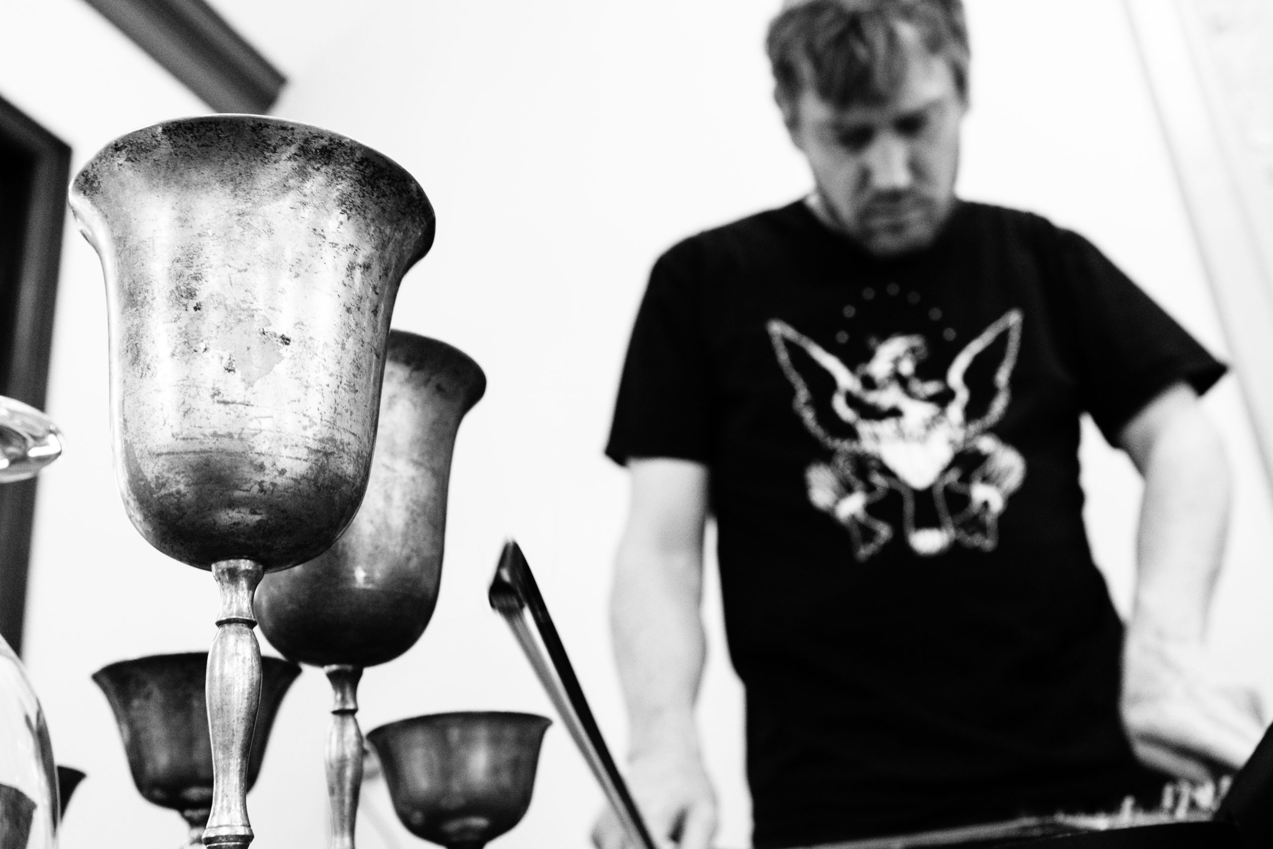 A black and white semi-focused shot of brass cups. A male musician with a dark black t-shirt stands behind, semi-blurred, playing music.