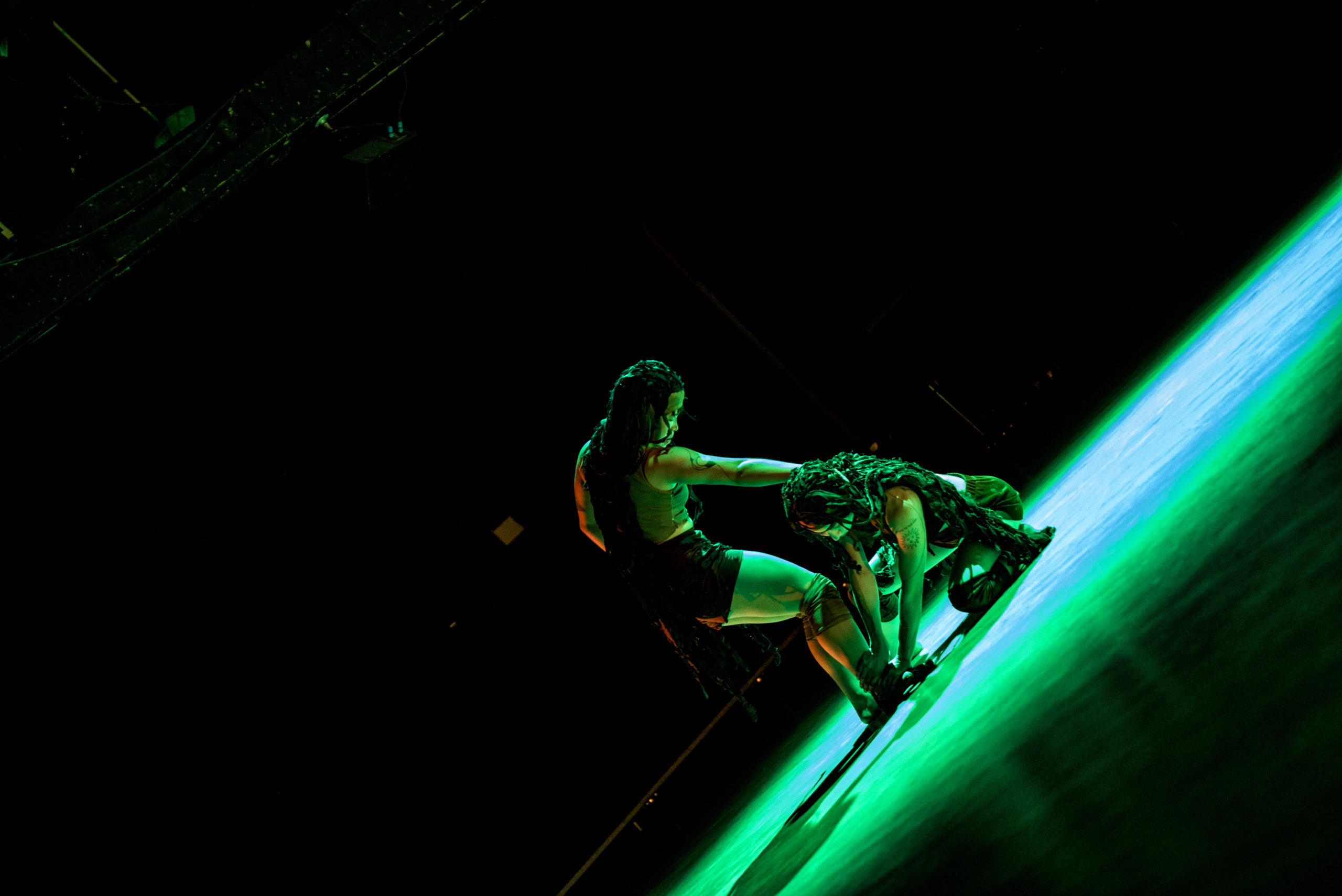 Two dancers, with long dark wigs covering their heads, perform on a wood-floored stage under green and blue lighting. One is crouched on the ground, the other stands over her arm wrapped around the back of the lower dancer’s neck.