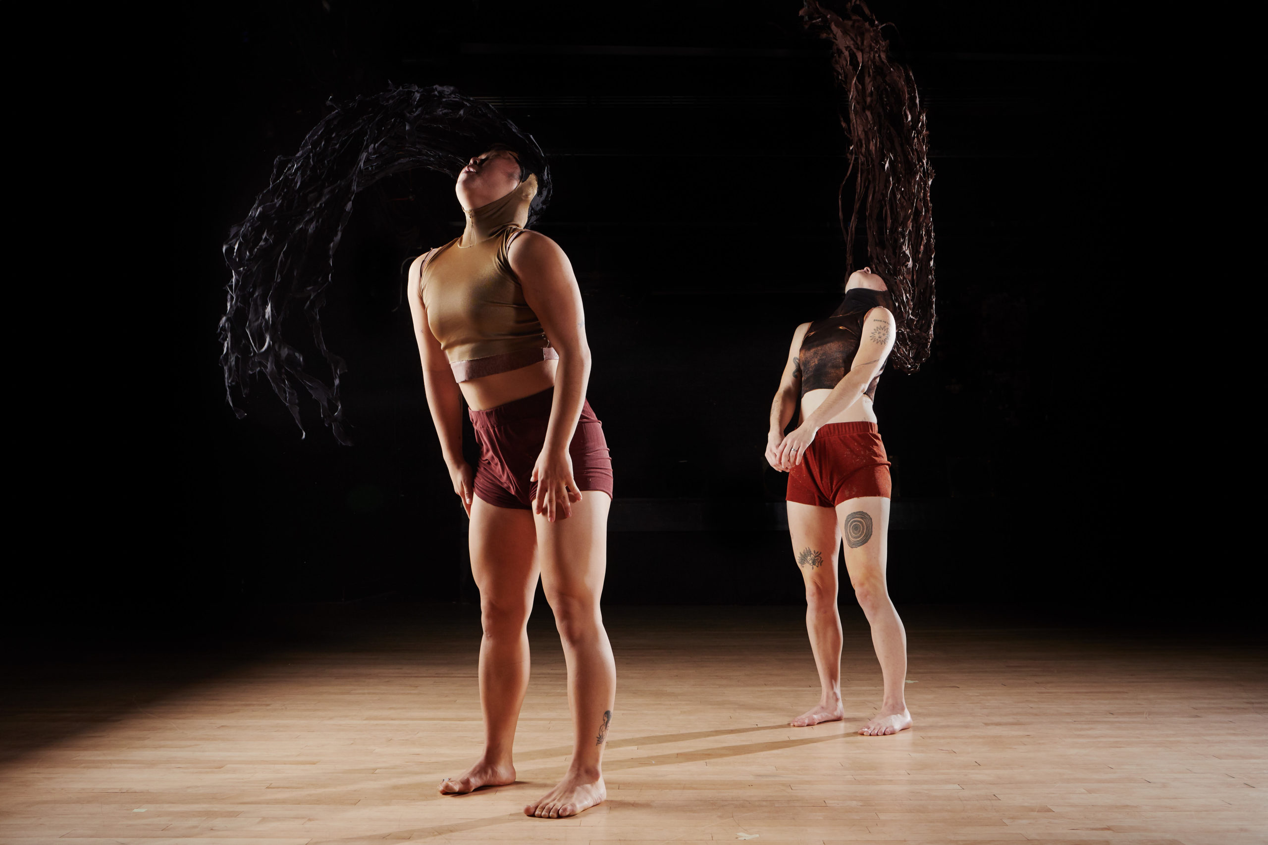 Two female bodied dancers wearing bronze cropped tops and tight, high wasted shorts toss their heads back and forth with long black wigs on their heads