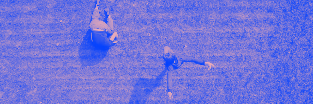 A wide, bird's eye view shot of two dancing bodies in the grass. One person is face dow in the grass, the other is upright, reaching their arms behind them. The whole image has been edited to look purple